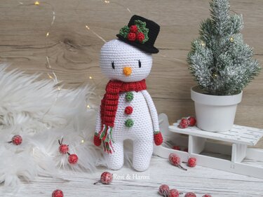 Gehäkelter Schneemann-Amigurumi mit schwarzem Zylinder, rotem Schal und bunten Knöpfen, umgeben von winterlicher Dekoration