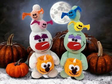 Two crocheted amigurumi figures, white and pale green, each holding a small crocheted pumpkin lantern with pumpkins and a full moon behind them.