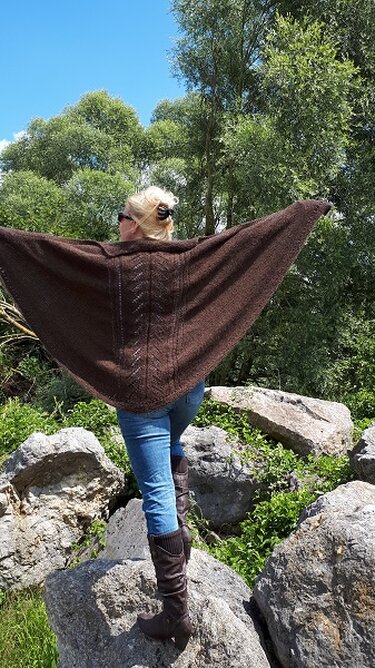 Person von hinten mit dunkelbraunem, gestricktem Tuch mit Strukturmuster, stehend auf Felsen vor Bäumen und blauem Himmel.