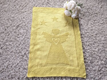 Pale yellow knitted baby blanket with a raised angel motif and three stars, textured border, laid on carpet with white flowers at the top right