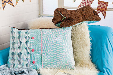 Türkises Dekokissen mit Blättermuster links und Blumenmuster rechts, rosa Zierknöpfe, auf weißer Fellauflage neben einem braunen Stoffhund.