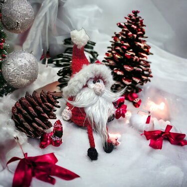 Gehäkelter Weihnachtswichtel mit rotem Hut und weißem Bart, umgeben von silbernen Kugeln, roten Schleifen und Tannenzapfen auf Deko-Schnee.