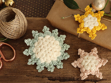 Three crocheted star and snowflake motifs in cream, sage green, pale pink and mustard on a wooden surface with twine and wrapped gift