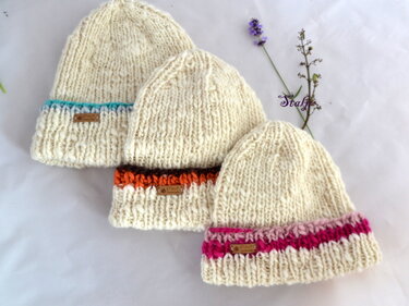 Drei cremefarbene Beanies in grobem Strick mit unterschiedlichen farbigen Umschlägen und kleinen Etiketten, auf weißer Fläche präsentiert.