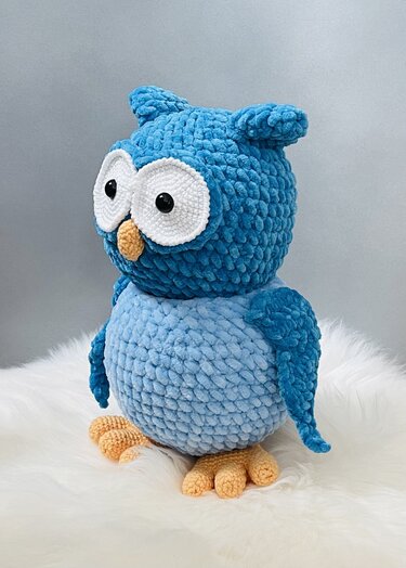 Gehäkelte blaue Amigurumi-Eule mit großen weißen Augen und orangefarbenem Schnabel auf weißem, flauschigem Untergrund.