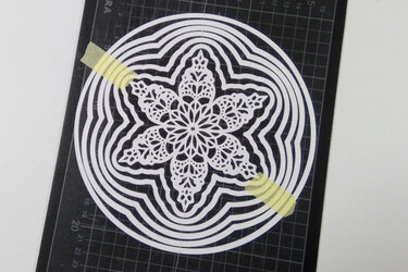 Weißes, rundes filigranes Mandala aus Papier auf schwarzer Raster-Schneidematte, mit zwei gelben Klebestreifen