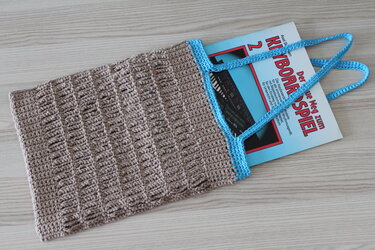 Beigefarbene gehäkelte Tasche mit hellblauem Rand und Henkeln auf Holz, mit einem Buch teilweise darin.