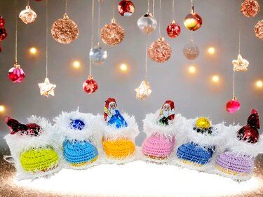 Row of colorful crocheted mini booties with white fuzzy trim holding foil-wrapped chocolates against a backdrop of hanging Christmas baubles and warm lights.