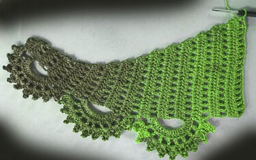 Halbfertiges gehäkeltes Dreieckstuch in Grün- und Olivtönen mit Bogenkante und Häkelnadel