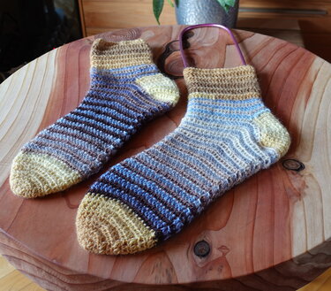 Zwei gehäkelte Socken mit horizontalen Streifen in Braun, Beige und Blau auf einer Holzplatte.