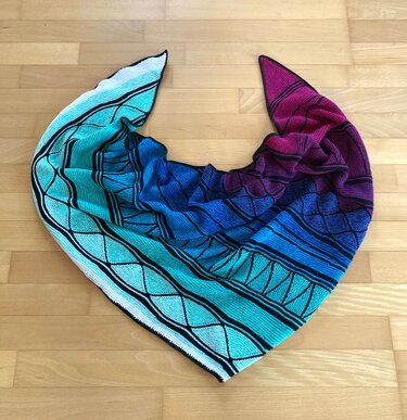 Gestricktes Dreieckstuch mit Farbverlauf von Türkis zu Magenta und schwarzem Linienmuster auf hellem Holzboden