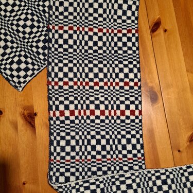 Gestrickter Schal mit Schachbrettmuster in Dunkelblau, Creme und Rot auf Holz.