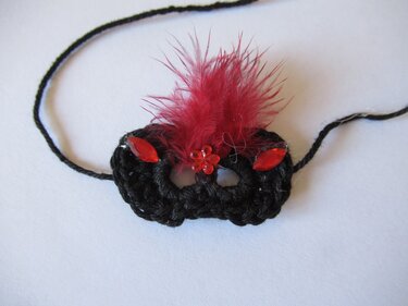 Schwarze gehäkelte Mini-Maske mit roten Federn und Schmucksteinen auf weißem Hintergrund.