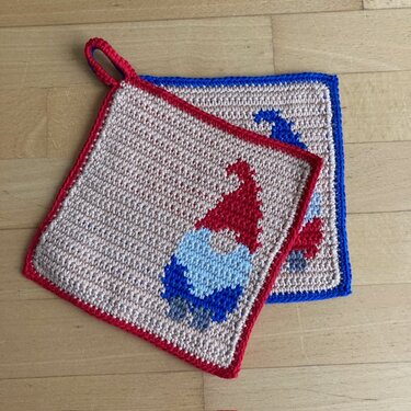 Zwei gehäkelte Topflappen mit Wichtelmotiven in Rot, Blau und Beige auf hellem Holzboden.
