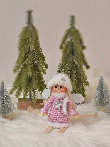 Gehäkeltes Amigurumi-Engelchen in rosa Kleid und weißer Mütze mit Mini-Skiern vor Deko-Tannen
