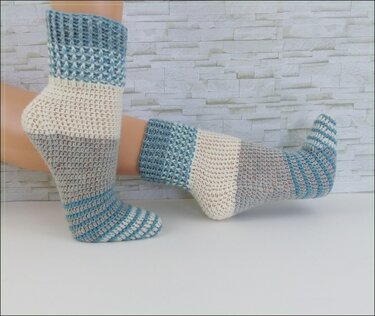Zwei Füße in handgehäkelten Socken mit Blockstreifen in Hellblau, Creme und Grau vor einer hellen Ziegelwand.