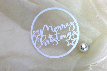 Weiß ausgeschnittener Kreis mit dem Schriftzug "Merry Christmas" und Sternmotiven auf hellem, strukturiertem Stoff; kleine silberne Kugel rechts.