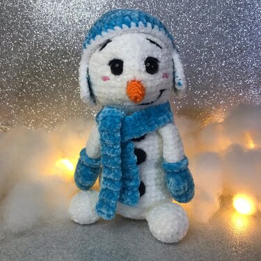 Gehäkelter Amigurumi
‑Schneemann in Weiß mit blauer Mütze, Schal und Fäustlingen, orange Karottennase und schwarzen Knöpfen vor glitzerndem Hintergrund mit Lichterkette.