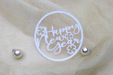 Weißer Kreis mit 'Happy New Year' Schriftzug und Kleeblättern auf hellem Stoff, zwei silberne Kugeln daneben.