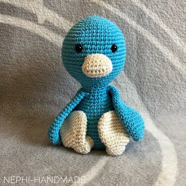 Sitzendes blaues Häkelvogel-Amigurumi mit cremefarbenem Schnabel und Füßen vor neutralem Hintergrund