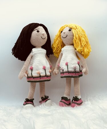Zwei gehäkelte Amigurumi-Puppen mit weißen Kleidern und rosa Blumen, eine mit schwarzem und eine mit gelbem Garnhaar