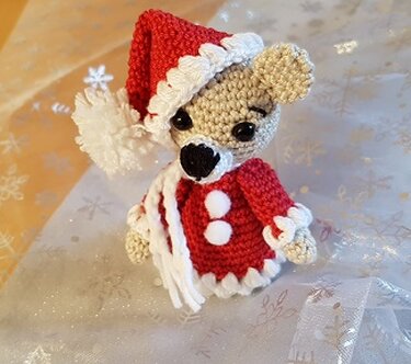 Gehäkelte Maus als Eierwärmer mit rotem Mantel, roter Zipfelmütze mit weißem Rand und weißem Schal auf einer Unterlage mit Sternmuster.