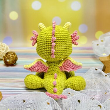 Back view of a lime green crocheted amigurumi dragon with pink wings and spines on star-patterned fabric.