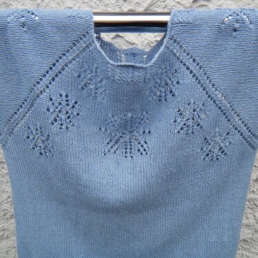 Light blue knitted sweater with snowflake eyelet motifs on the yoke and raglan shoulders, hung on a hanger.