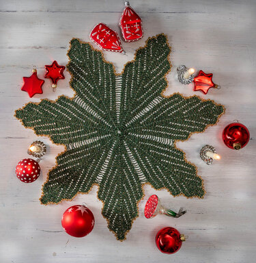 Sternförmiges grünes Häkeldeckchen mit goldener Kante auf hellem Holz, umgeben von roten Christbaumkugeln und kleinen Kerzen.