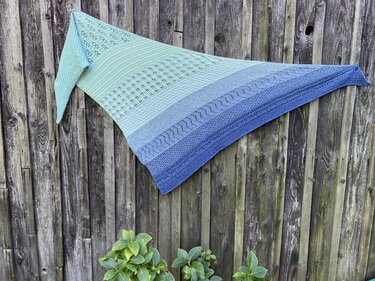 Gestricktes Dreieckstuch mit Türkis-Blau-Farbverlauf und verschiedenen Musterabschnitten an einem Holzzaun