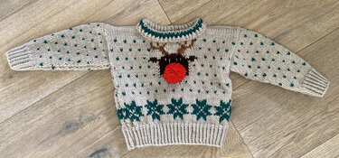 Beigefarbener handgestrickter Baby-Pullover mit Rentiermotiv und roter Pompom-Nase auf Holzboden.