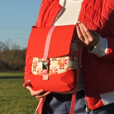 Person in roter Steppjacke hält eine rote Stofftasche mit floralem Muster und Schnallenverschluss im Freien.