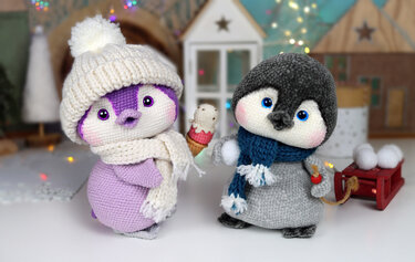Two crocheted penguin amigurumi — lavender with cream hat and scarf holding an ice-cream prop, and gray with blue scarf beside a small red sled with snowballs