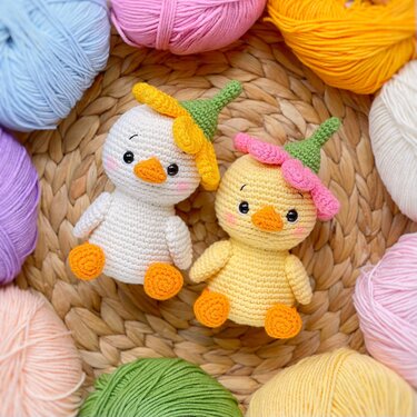 Two crocheted amigurumi chicks with orange beaks and feet, wearing small flower-style hats, on a woven mat surrounded by colorful yarn balls.
