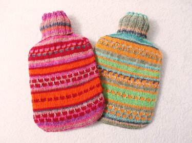 Zwei bunte gestrickte Wärmflaschenhüllen in Rot-Orange und Grün-Orange auf hellem Hintergrund.