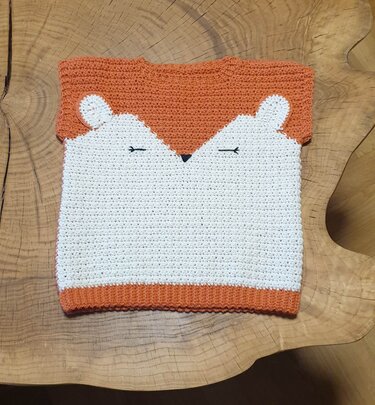 Gehäkelter Baby- und Kinderpullover in Orange und Weiß mit stilisiertem Fuchsgesicht, flach auf Holzuntergrund ausgelegt