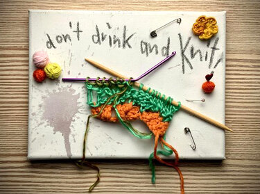 Leinwand mit Schrift 'don't drink and knit', Holzstricknadeln, violette Häkelnadel und grün-orangem Garn