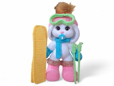 Gehäkelter weißer Amigurumi-Hase mit gelbem Snowboard, grünen Skistöcken, grüner Skibrille und rosa Mütze