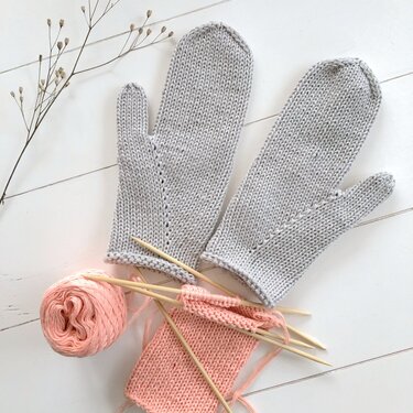 Hellgraue gestrickte Fäustlinge auf weißem Holz, dazu rosafarbene Wolle, Stricknadeln und ein angefangenes Strickprojekt.