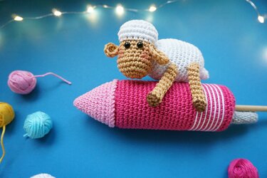Weißes gehäkeltes Amigurumi‑Schaf mit beigefarbenem Kopf und Beinen auf einer rosa‑pink gehäkelten Rakete, hellblaue Unterlage, Garnknäuel und Lichterkette im Hintergrund.