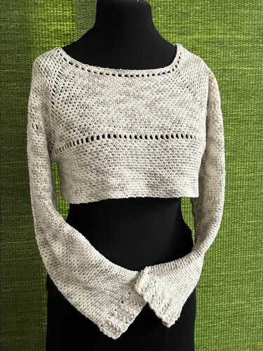 Gehäkelter hellbeiger, kurz geschnittener Damenpullover mit langen Ärmeln auf schwarzer Schaufensterpuppe vor grünem Hintergrund