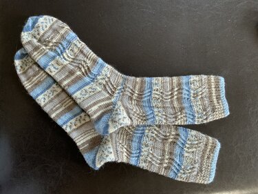 Handgestricktes Paar Socken in Blau-, Grau- und Hellgrautönen mit Streifen- und Strukturmuster auf dunklem Hintergrund.