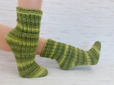 Zwei handgehäkelte grüne Socken mit Rippen- und Strukturmuster an ausgestellten Füßen vor hellem Hintergrund.