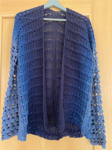 Gehäkelte blaue Damenjacke mit offenem Vorderteil an einem Kleiderbügel, sichtbar Loch- und Rippenmuster