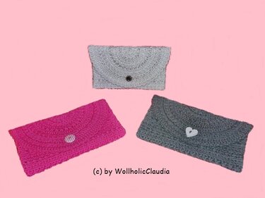 Drei gehäkelte Umschlag-Clutches in Pink, Hellgrau und Dunkelgrau mit Knopfverschluss auf rosa Hintergrund