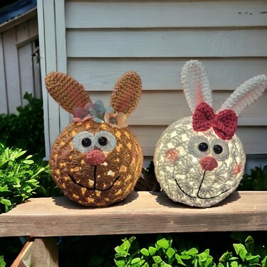 Zwei gehäkelte Hasenköpfe als Lichter, braun mit Blumen und weiß mit rosa Schleife, auf einer Holzleiste.