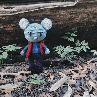 Gehäkelte hellblaue Amigurumi‑Maus mit roter Weste vor Baumstamm