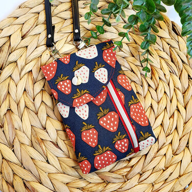 Dunkelblaue Stofftasche mit rotem und weißem Erdbeermuster, Umschlagklappe mit Druckknopf und Reißverschlussfach auf geflochtener Unterlage mit grünem Blattwerk.