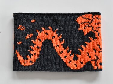 Rectangular knitted scarf or cowl with a bold orange flame-like pattern on a dark gray background.
