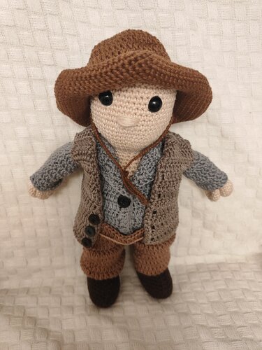 Gehäkelte Amigurumi-Puppe mit braunem Hut, grauem Oberteil, brauner Weste, Hose und dunklen Stiefeln vor hellem Stoffhintergrund